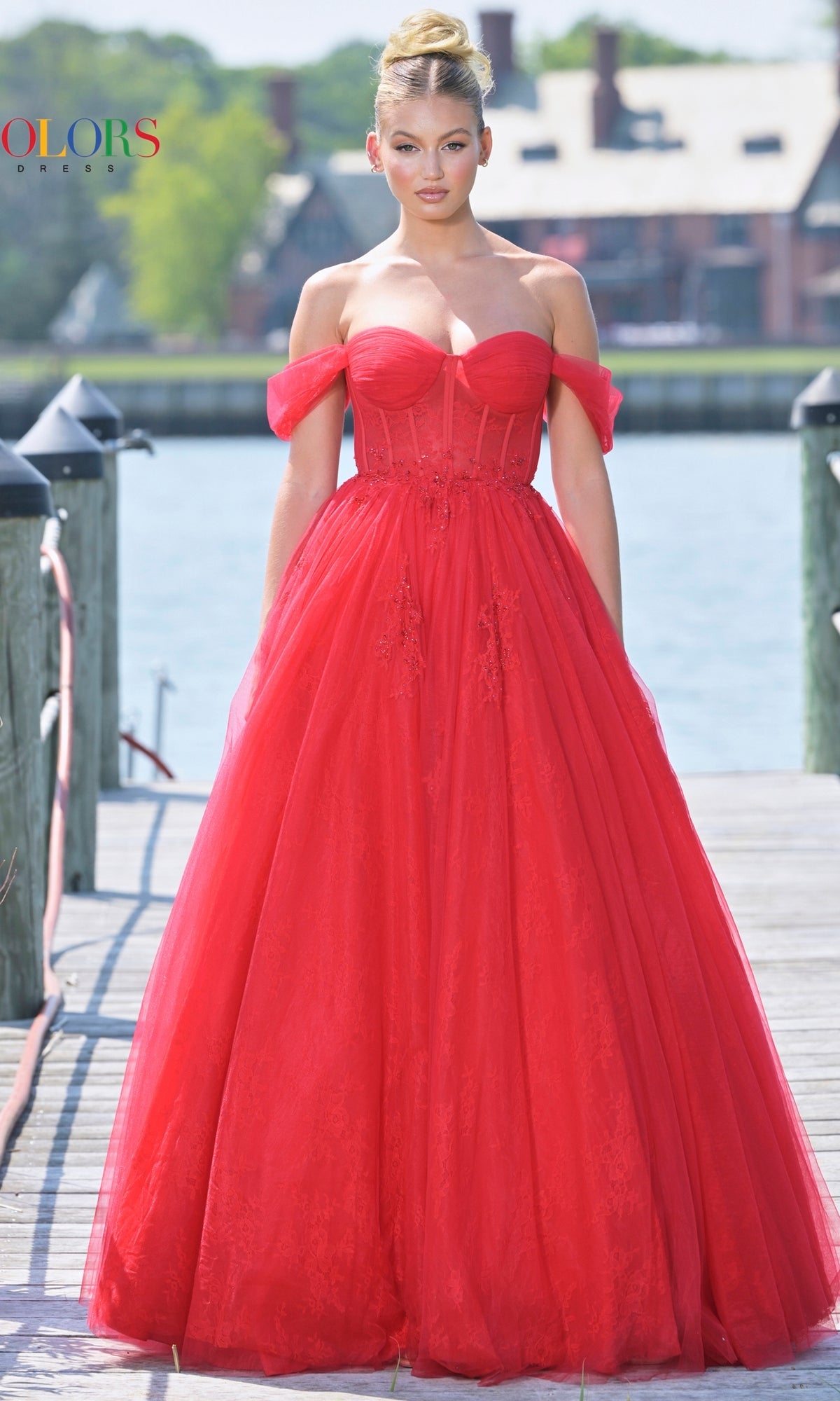 A woman in the Long Sweetheart Prom Ball Gown: Colors Dress 3240, featuring a strapless red fitted bodice and tulle skirt, stands on a dock by the water.